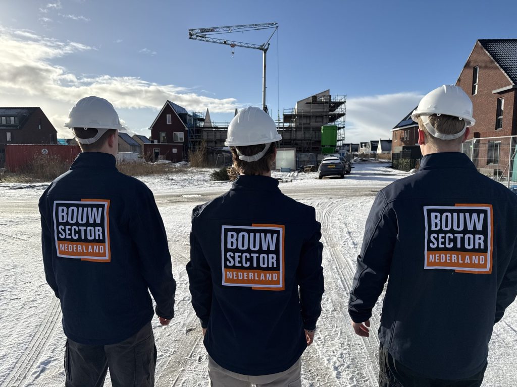 Drie medewerkers van Bouwsector Nederland met witte helm en donkere werkjas kijken naar huizen in aanbouw in de sneeuw